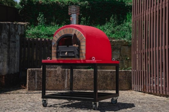 Un four à bois extérieur de couleur rouge vif, mobile sur roulettes, installé sur une terrasse en pierre devant un mur végétal
